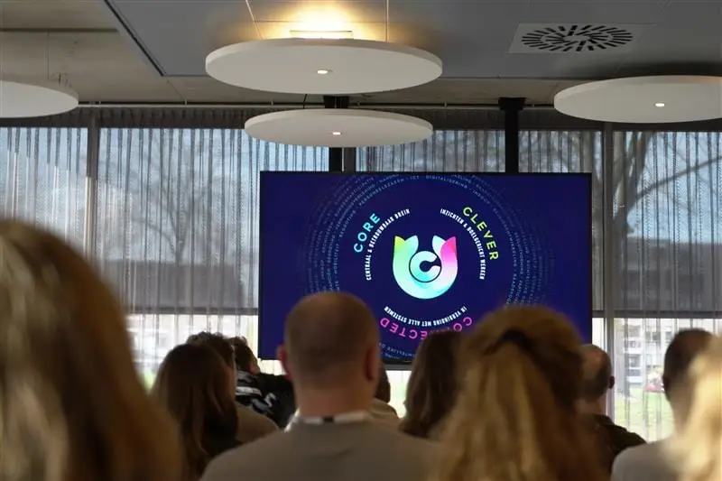Zaal met aanwezigen die kijken naar een presentatiescherm met het logo en de drie kernwaarden van Corsa: Core, Clever en Connected.