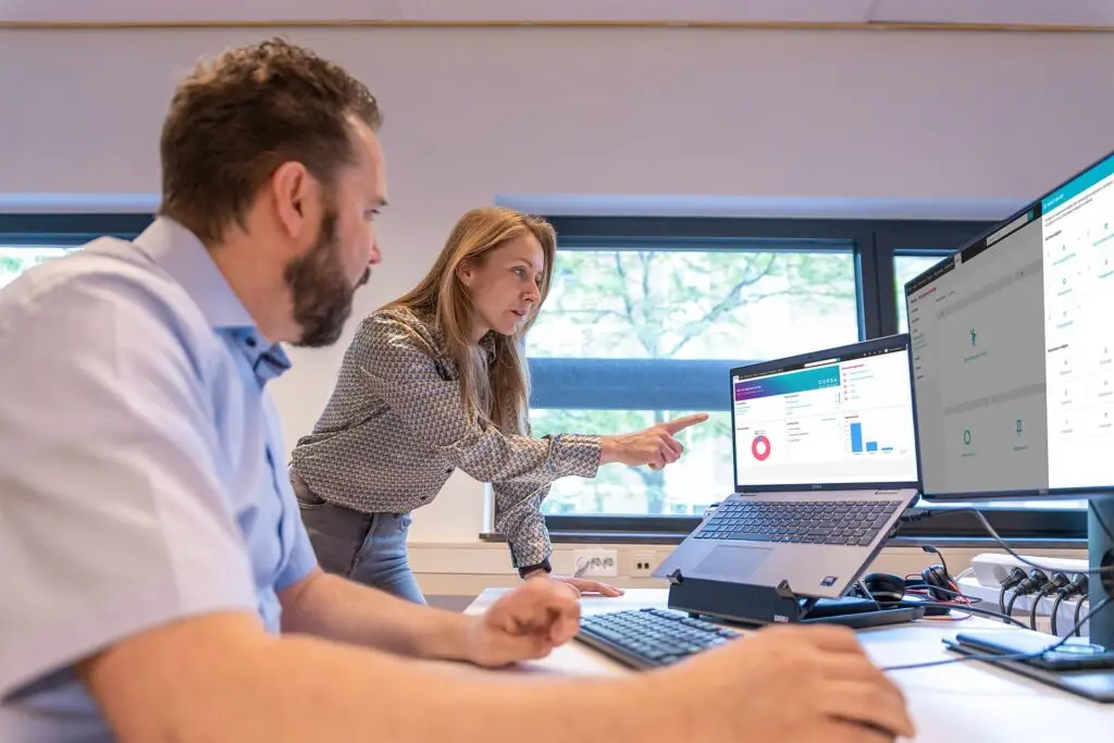 jonge vrouw wijst naar een computerscherm. Mannelijke collega kijkt toe.