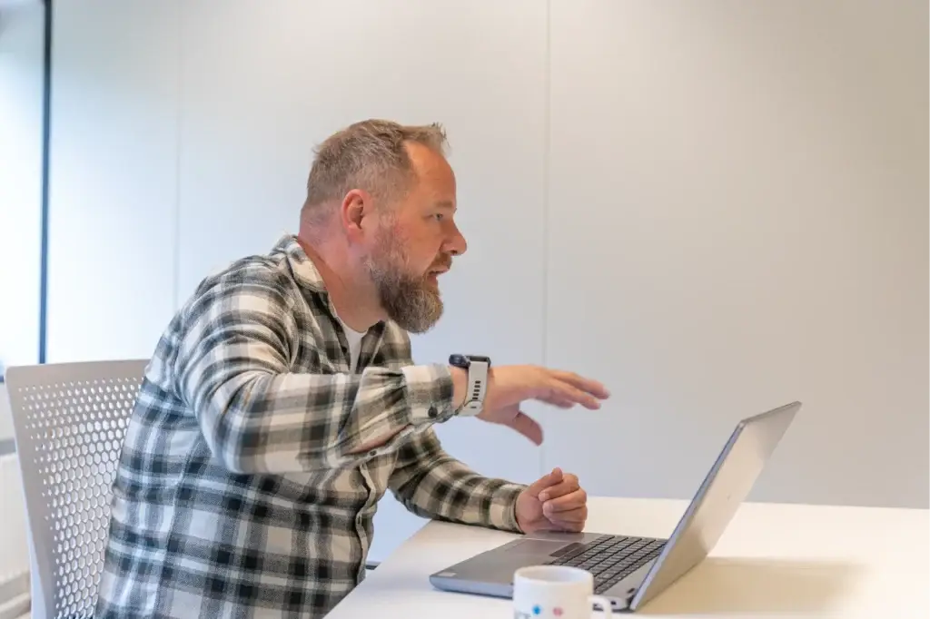 Man in geruit overhemd wijst geconcentreerd naar het scherm van zijn laptop tijdens zijn werk.