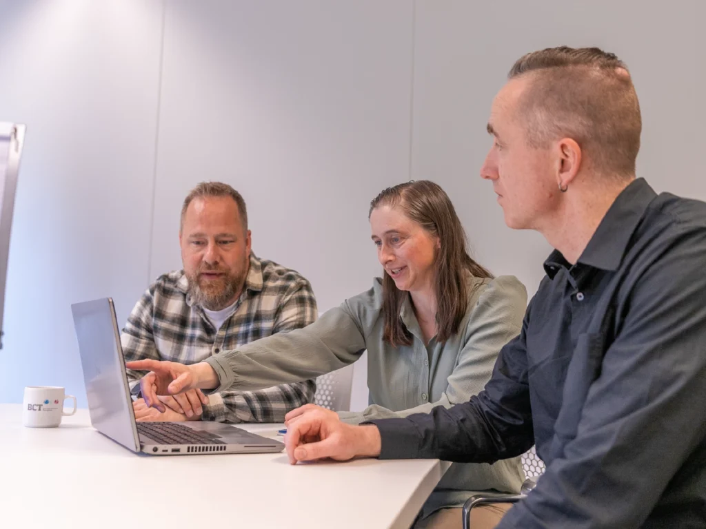 Drie collega's overleggen samen aan een tafel. Een vrouw wijst iets aan op een laptop, terwijl twee mannen meekijken.