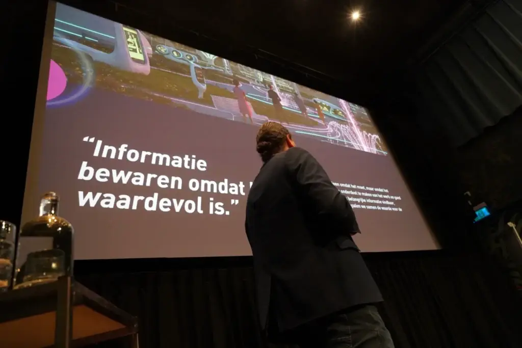 Spreker op een evenement voor een groot projectiescherm met de quote: "Informatie bewaren omdat het waardevol is."