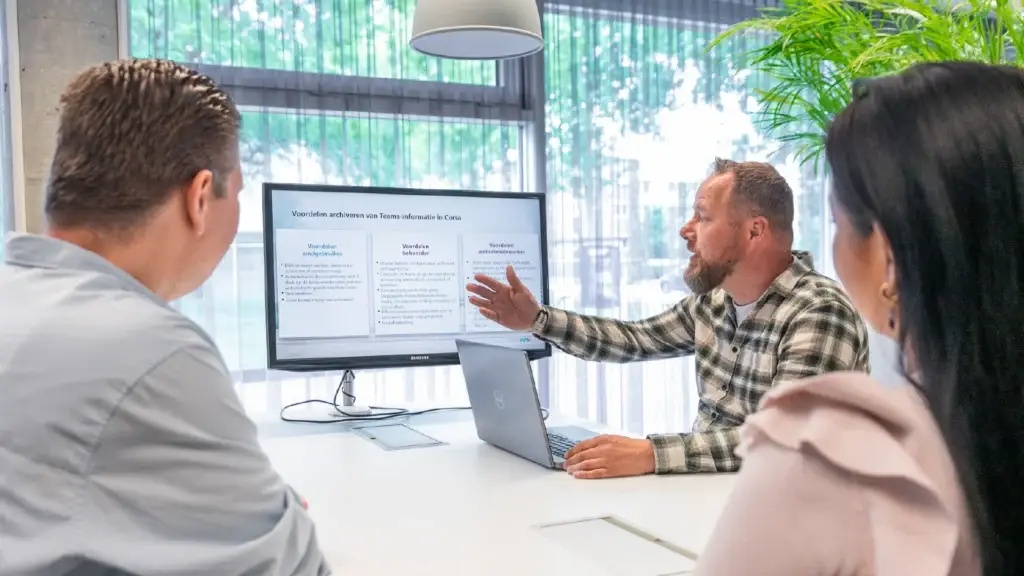 Consultant legt twee collega's uit aan een vergadertafel, wijzend naar een scherm met een presentatie en beantwoordt vragen over in Corsa.