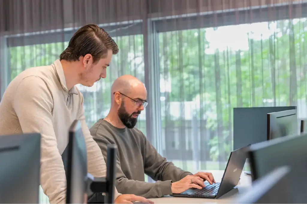 Twee collega's kijken samen naar een laptopscherm in een kantooromgeving.