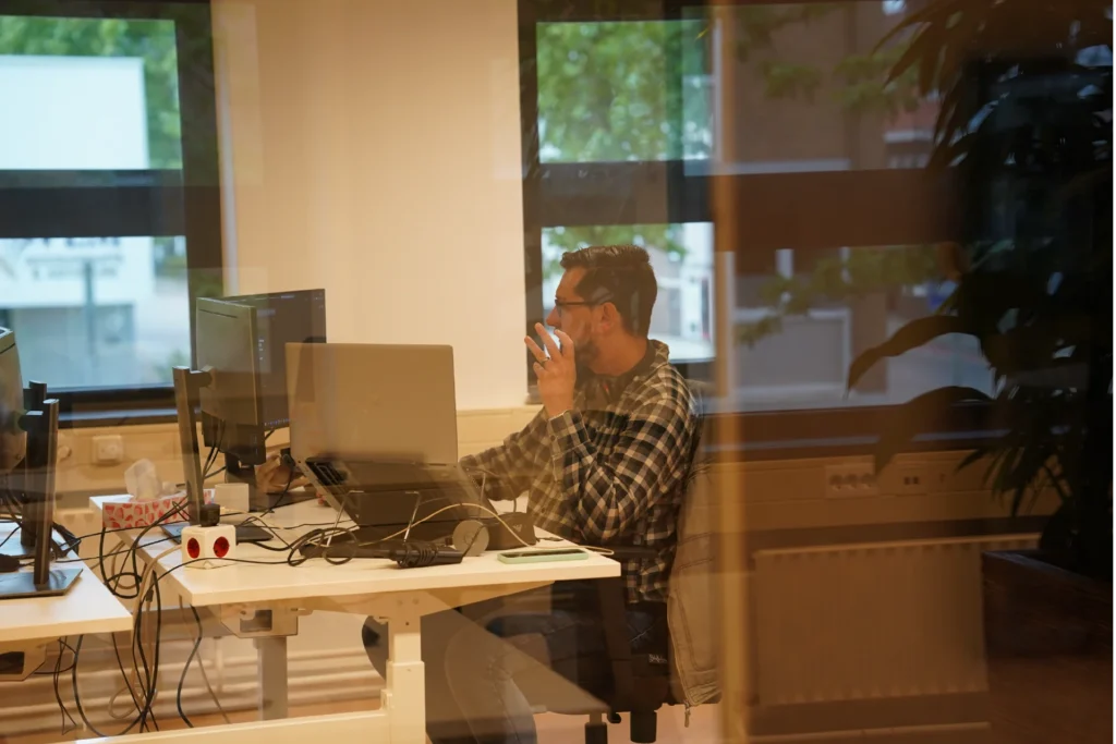 Medewerker achter een werkplek met meerdere schermen, gefotografeerd door een glazen wand.