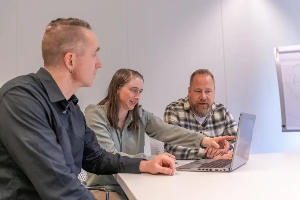 Drie personen bekijken samen een laptop, waarbij één persoon iets aanwijst op het scherm.