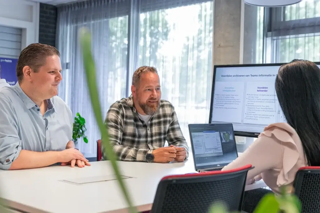 Drie collega's in overleg aan een vergadertafel, met op de achtergrond een scherm met een Corsa-presentatie.