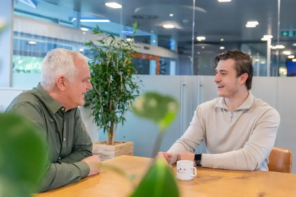 Twee collega's in gesprek aan een tafel met koffie en planten op de achtergrond