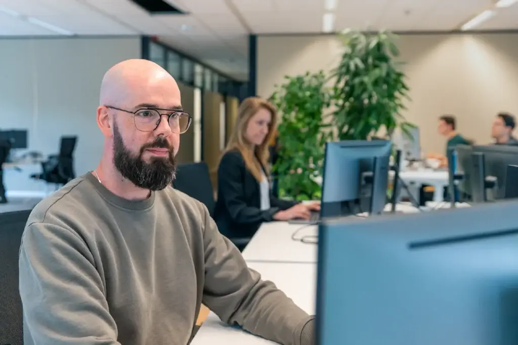 Medewerker werkt geconcentreerd achter het beeldscherm, collega's zijn op de achtergrond te zien