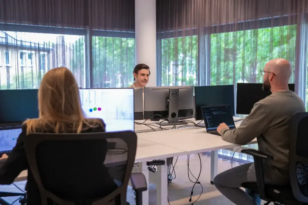 Drie collega's werken aan bureaus met meerdere beeldschermen in een licht kantoor